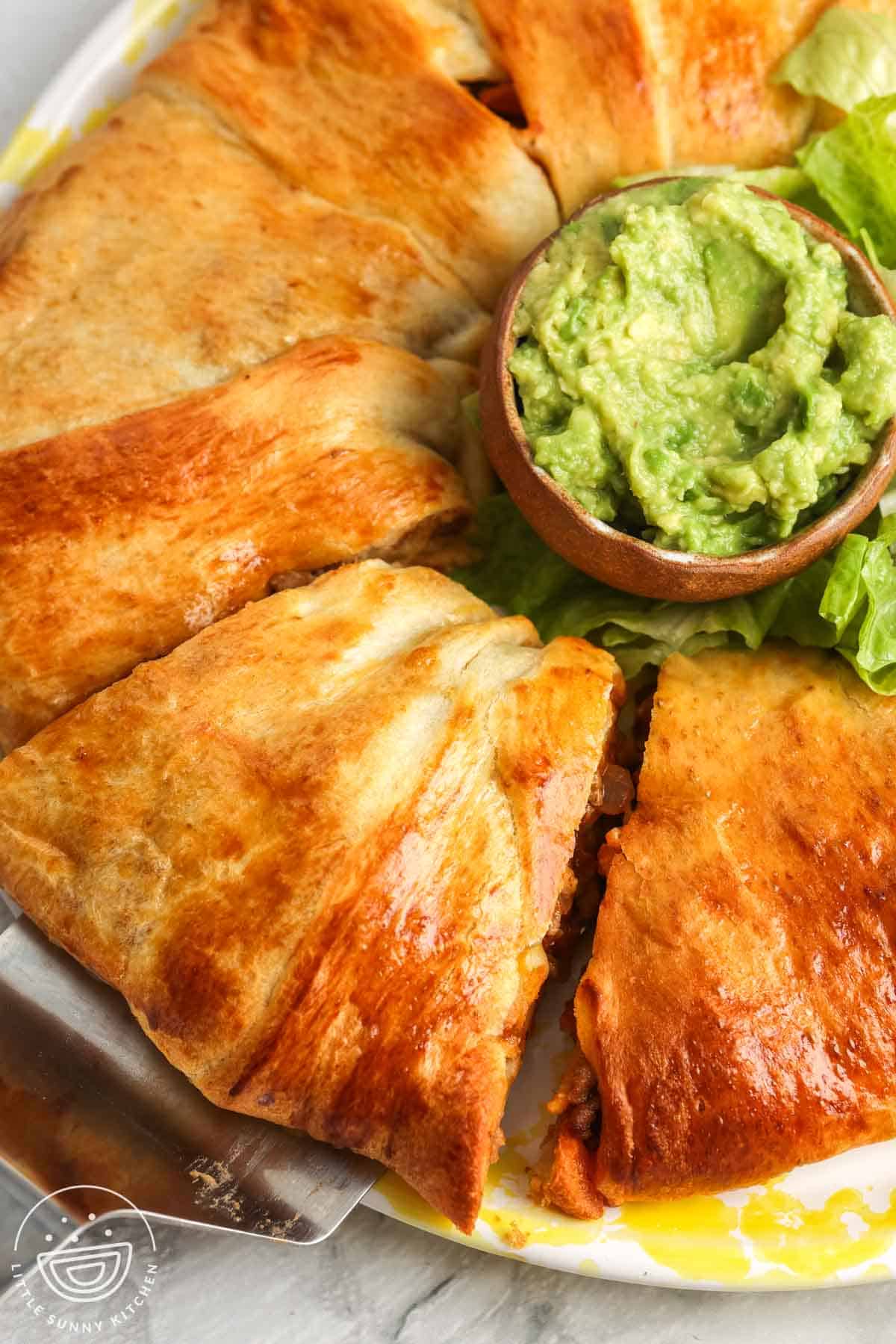 Overhead view of a baked taco ring made with crescent roll dough, sliced into sections and served on a plate with a small bowl of guacamole and lettuce on the side, showing the golden browned crust and filled center