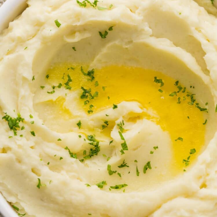 Overhead and close up shot of creamy mashed potatoes served in a white bowl with melted butter and fresh herbs