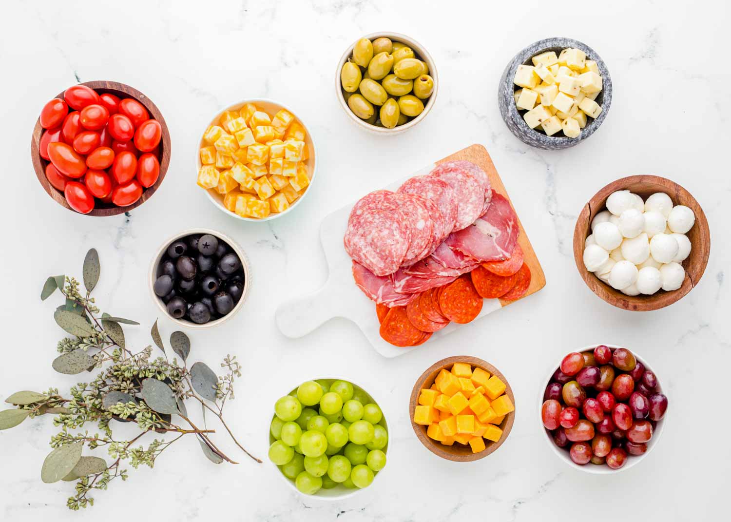 ingredients needed to make a charcuterie board including cheeses, meats, grapes, and cherry tomatoes.