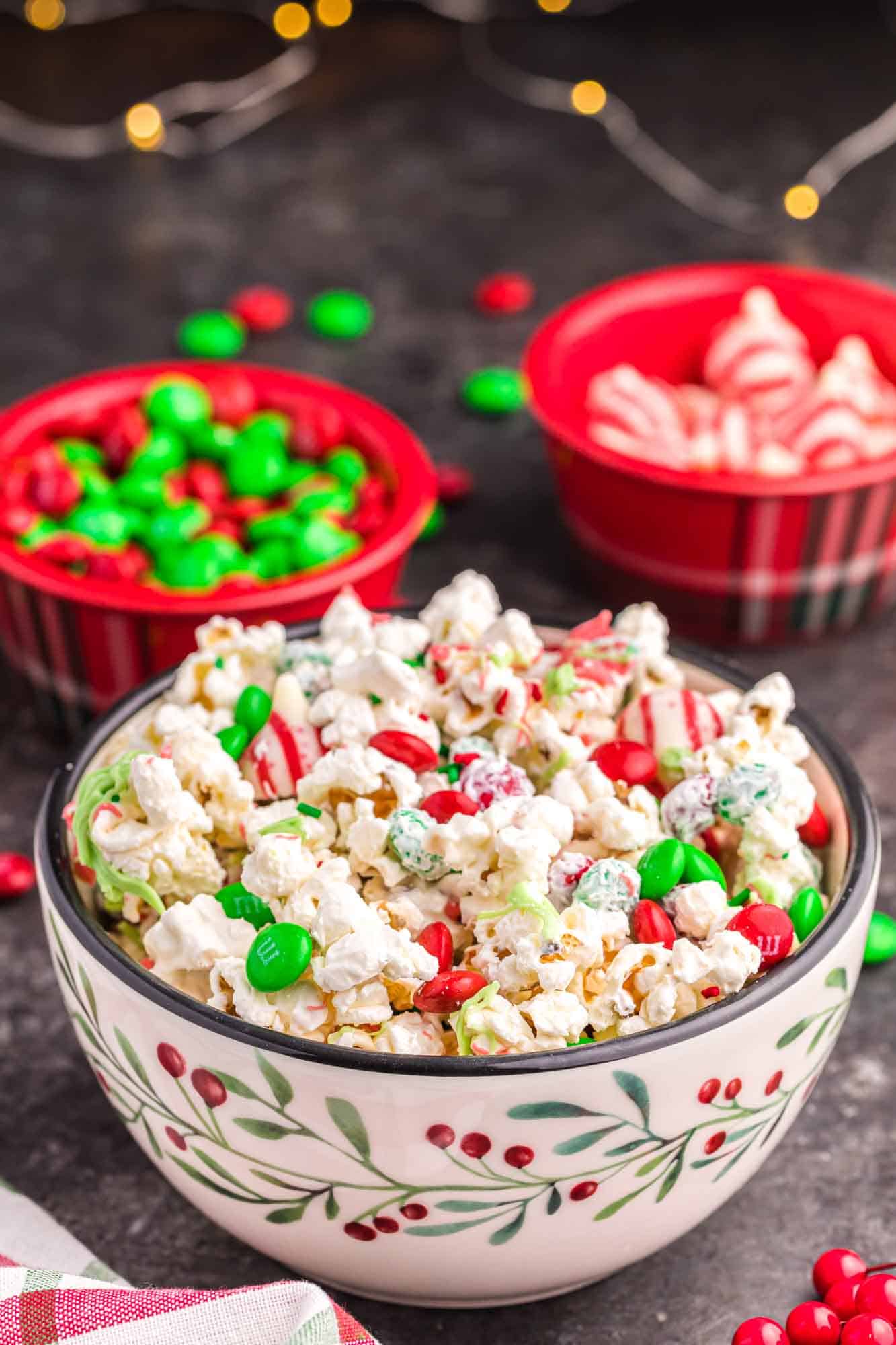 A bowl of christmas popcorn with christmas light in the background and small bowls of treats