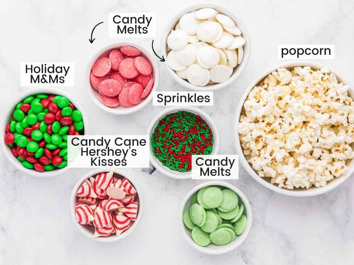 Ingredients needed to make Christmas Popcorn including popped popcorn, melts, M&Ms, sprinkles, Candy cane hershey's kisses.