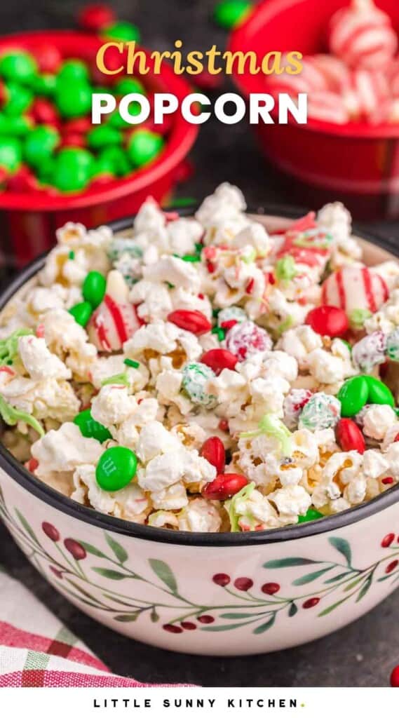 Christmas popcorn served in a festive bowl, with holiday M&Ms and candy cane hersheys in the background. And overlay text that says "Christmas Popcorn"