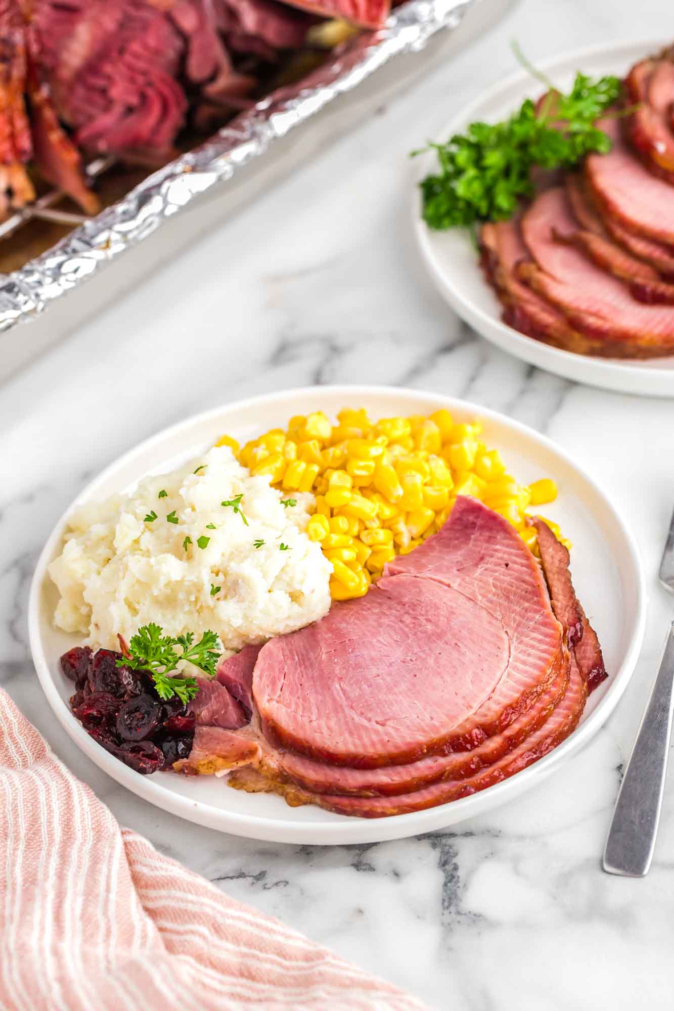 Sliced ham served on a plate with a side of mashed potatoes and corn.