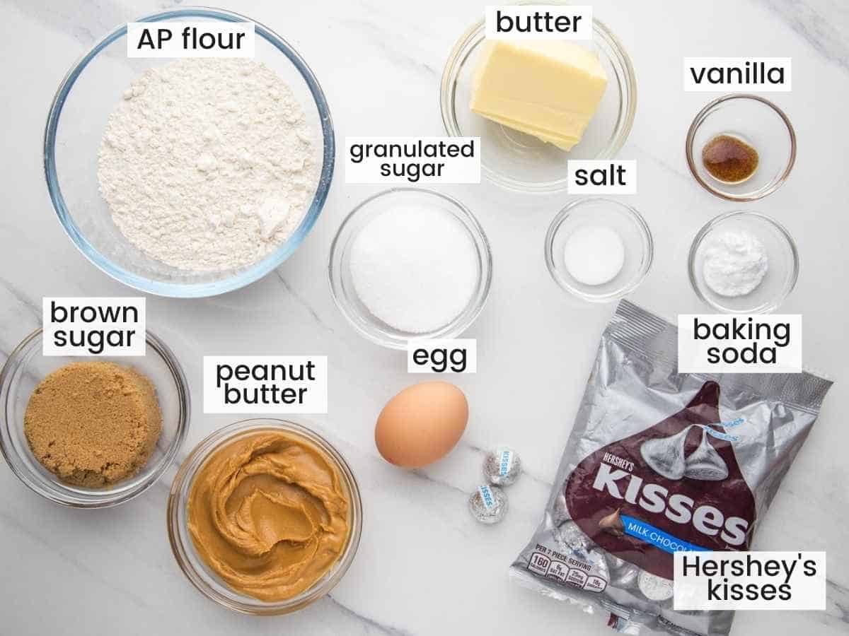 Ingredients needed to make peanut butter blossoms including flour, sugars, egg, butter, peanut butter, baking soda, salt, vanilla, and hersheys kisses.