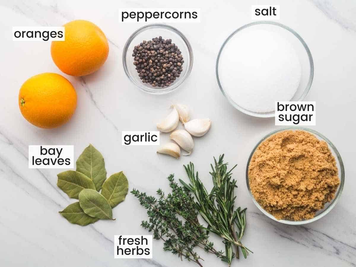 Ingredients needed to make turkey brine including oranges, salt, brown sugar, bay leaves, garlic, peppercorns, and fresh herbs.