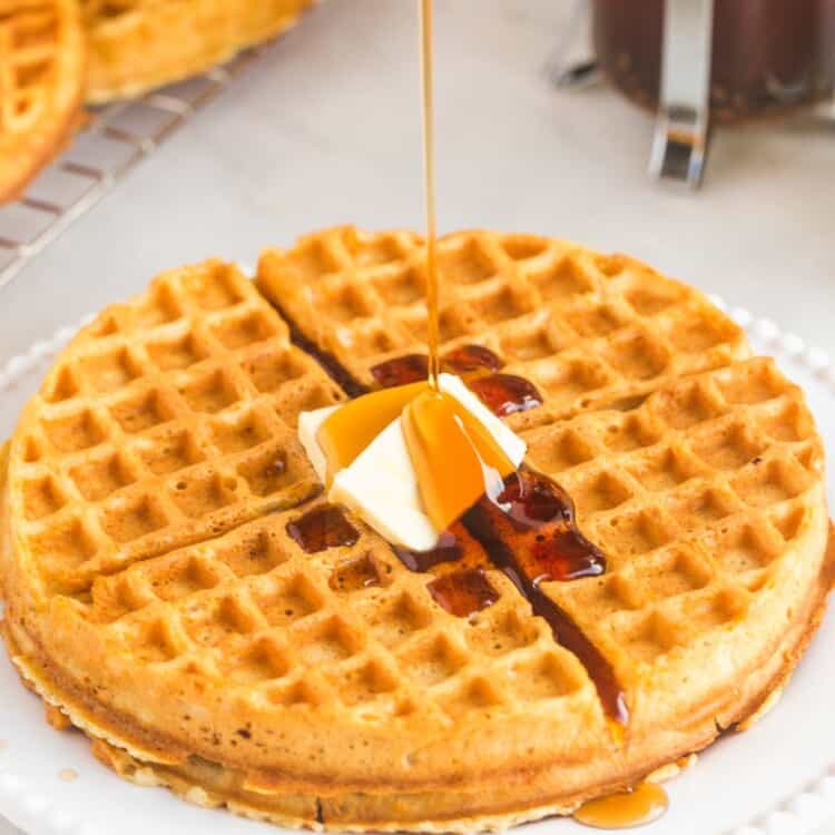 A round homemade waffle served on a white plate with butter and maple syrup, with french press coffee in the background.