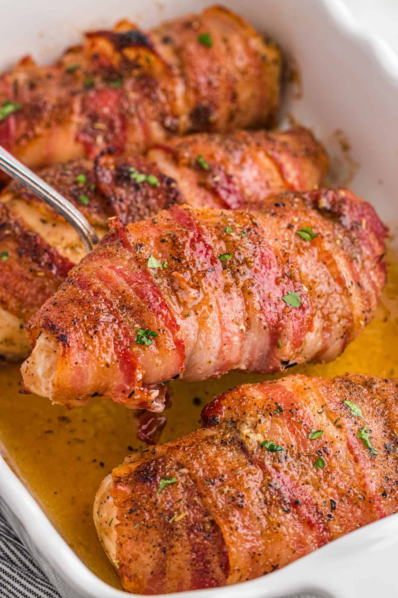 Taking one bacon wrapped chicken breast from a white baking dish