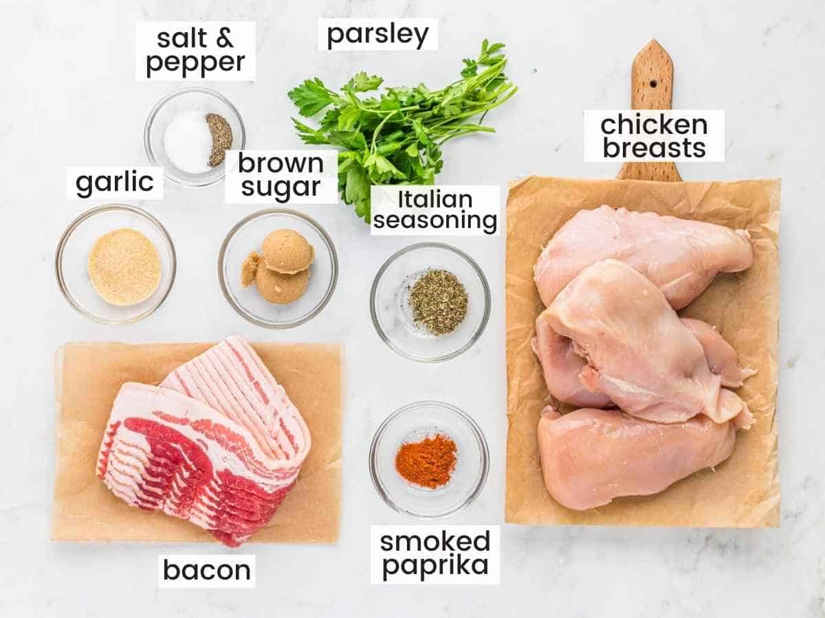 Ingredients needed for making bacon wrapped chicken breasts including chicken breasts, bacon, seasonings, and parsley.
