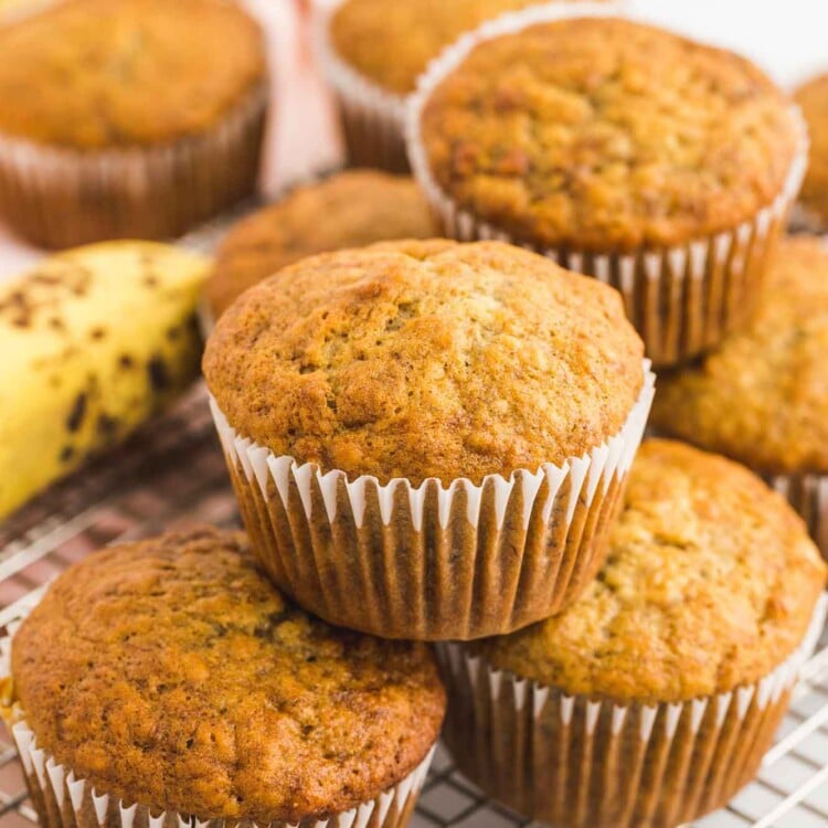 Stacked banana muffins placed on a wire rack, with ripe bananas in the background.