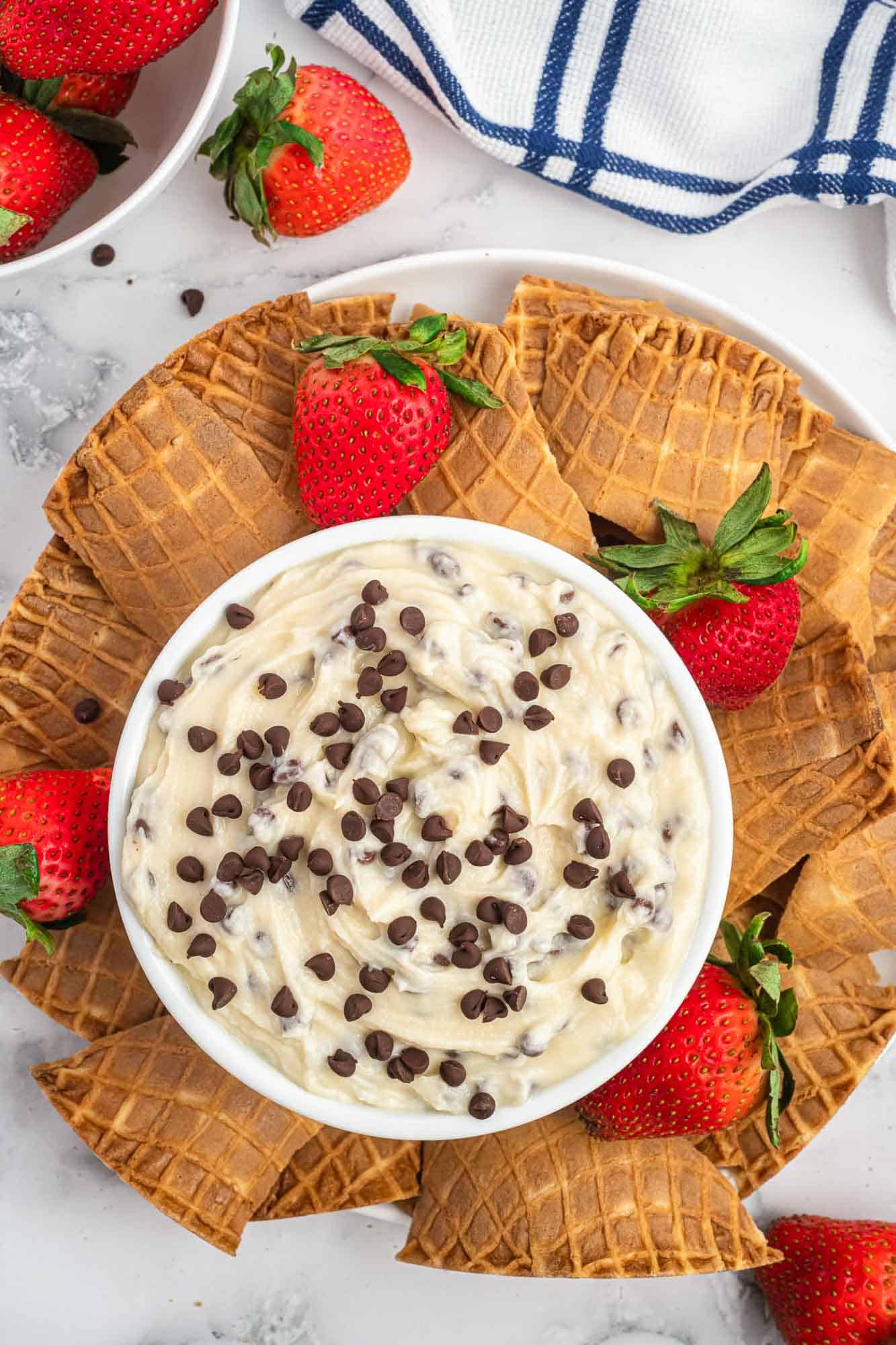 Overhead shot of a bowl with cannoli dip with mini chocolate chips, broken up cone chips, and fresh strawberries.