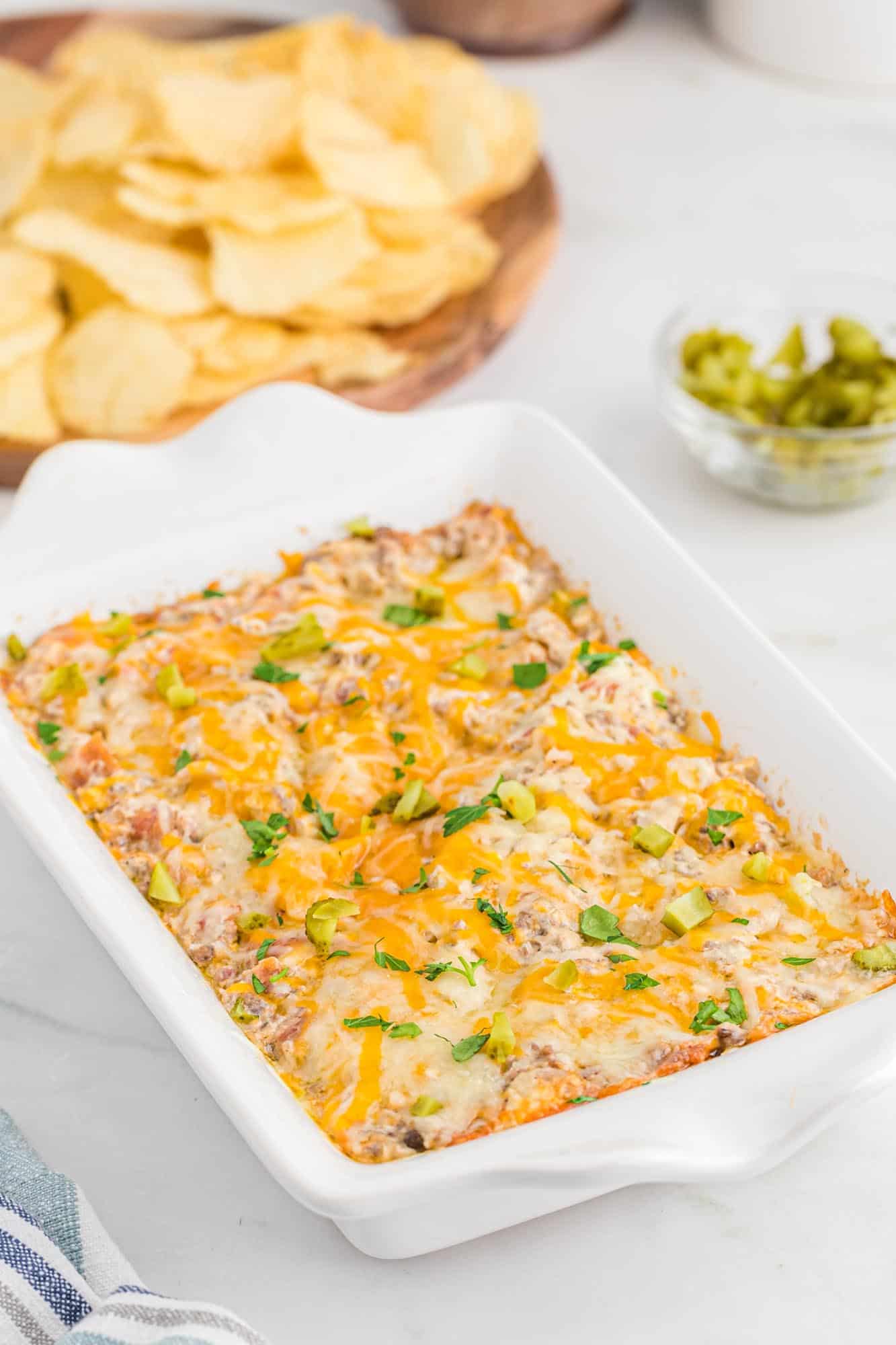 Cheeseburger dip in a small white baking dish, covered with melted cheese. And potato chips in the background
