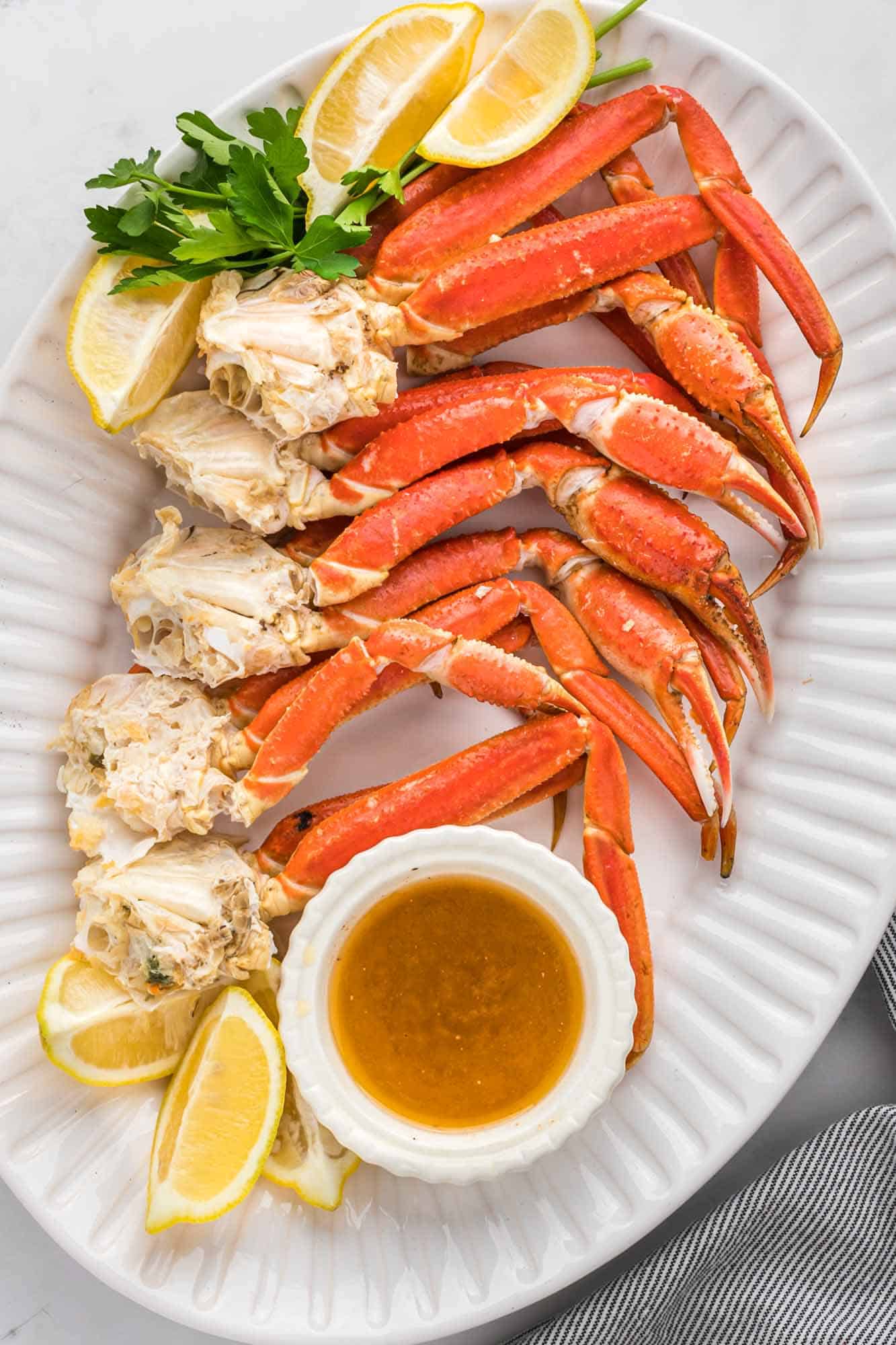 Overhead shot of crab legs served on a large oval white platter with butter dipping sauce and lemon wedges.