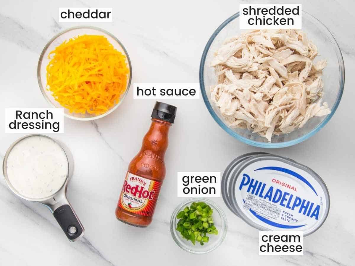 ingredients needed to make slow cooker buffalo chicken dip including shredded chicken, hot sauce, ranch, cheddar, cream cheese and green onions.
