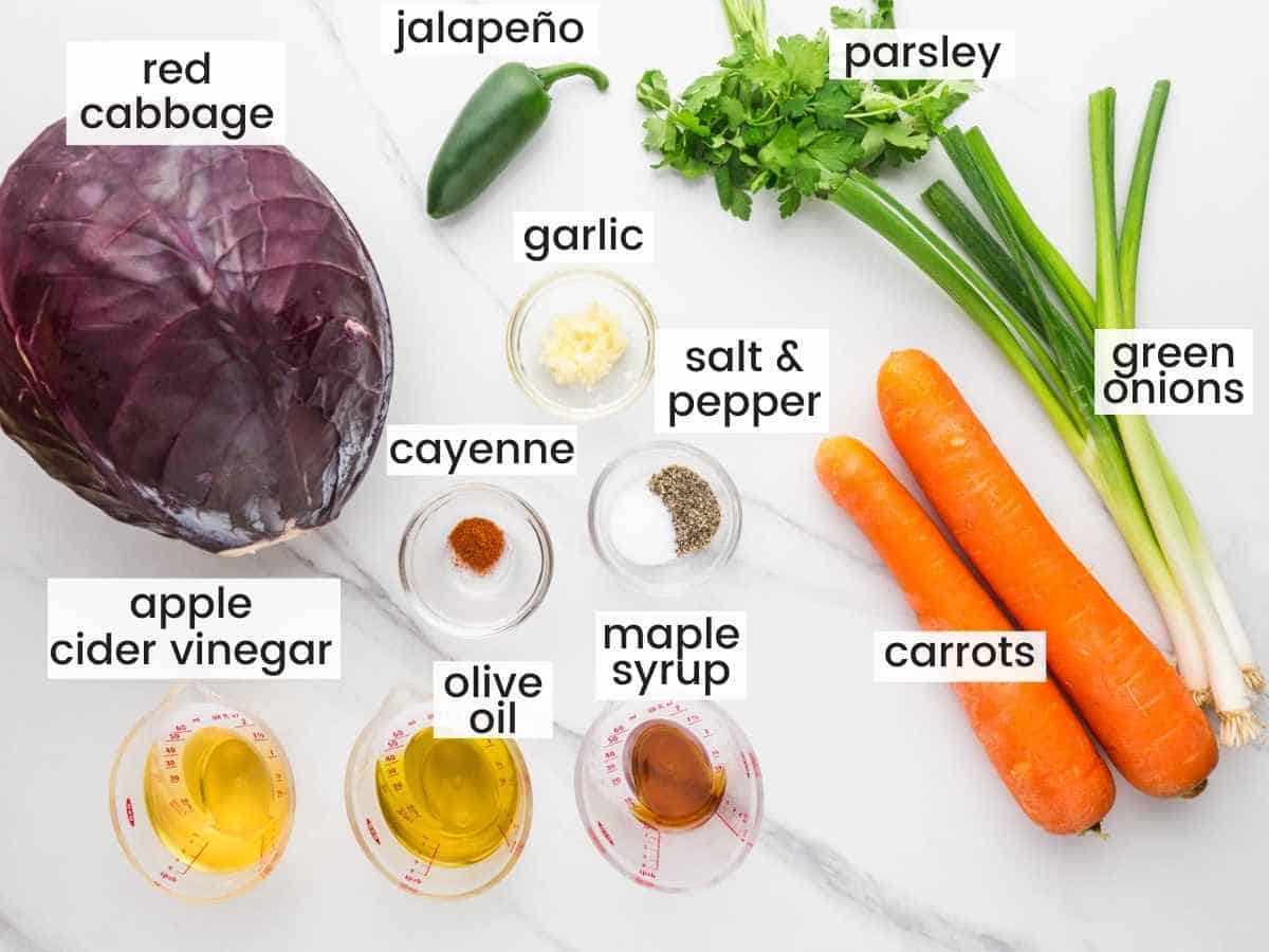 Ingredients needed to make red cabbage slaw including red cabbage, carrots, jalapeno, parsley, green onions, garlic, maple syrup, apple cider vinegar, olive oil, cayenne, salt and pepper.