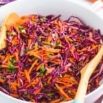 Red cabbage slaw served in a large white salad bowl, with wooden serving spoons.