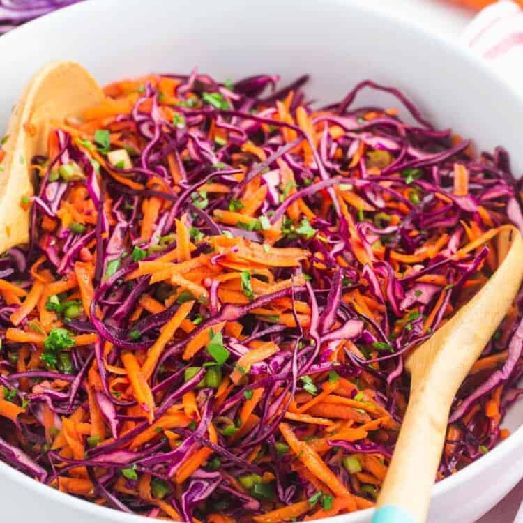 Red cabbage slaw served in a large white salad bowl, with wooden serving spoons.
