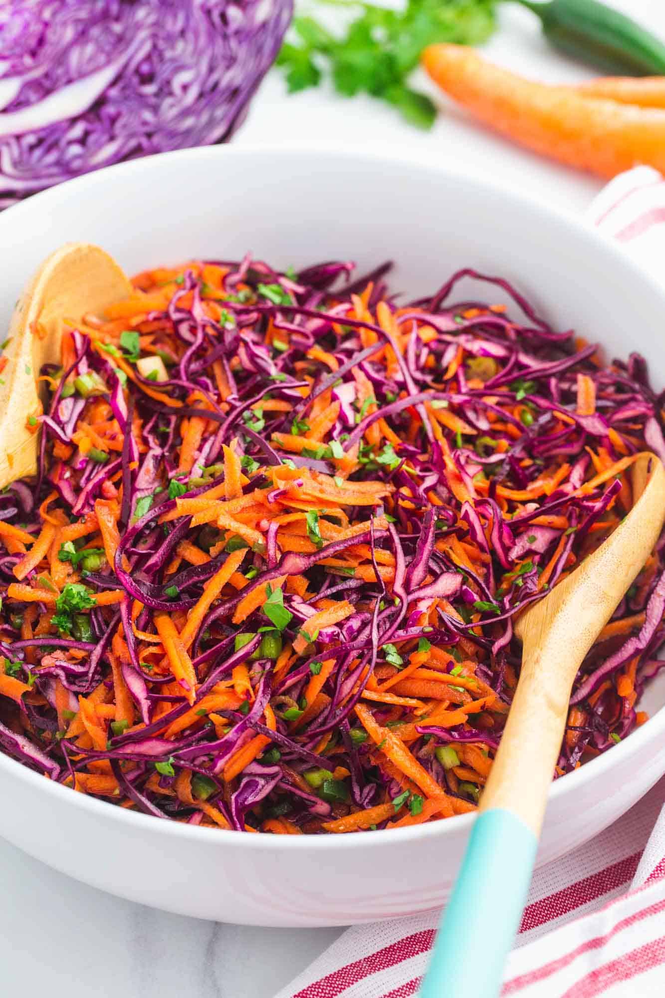 Red cabbage slaw served in a large white salad bowl, with wooden serving spoons.