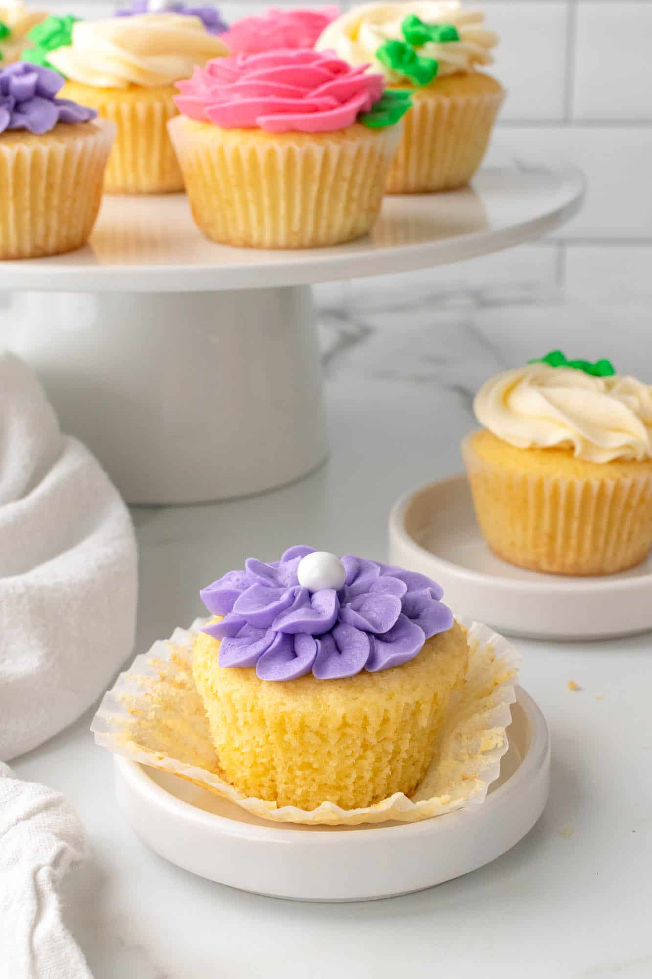 Purple flower cupcake on a small plate, and a cake stand with more cupcakes in the background