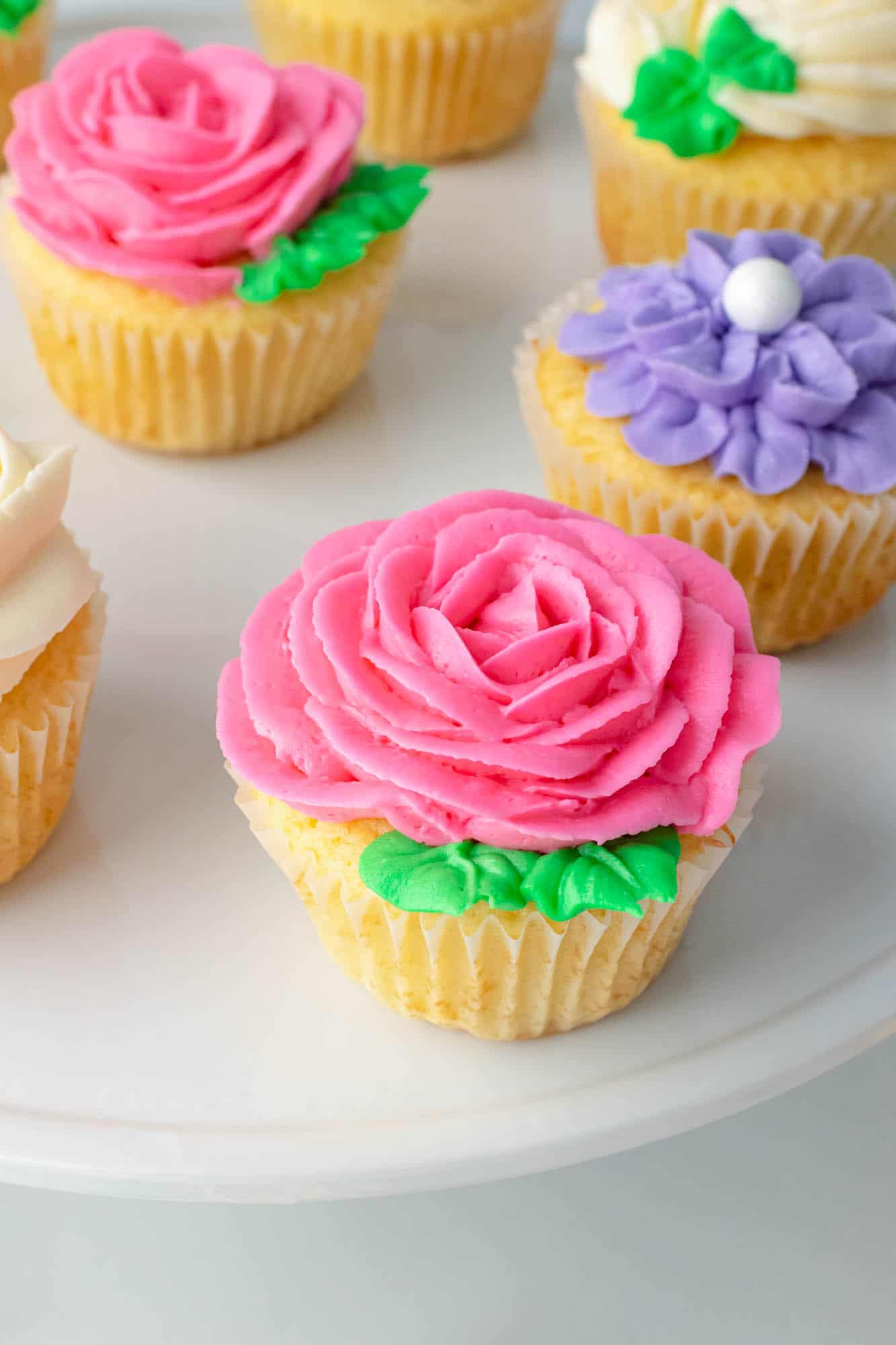 Cupcake with a buttercream rose piped onto it, with green leaves on the side. And more flower cupcakes in the background.