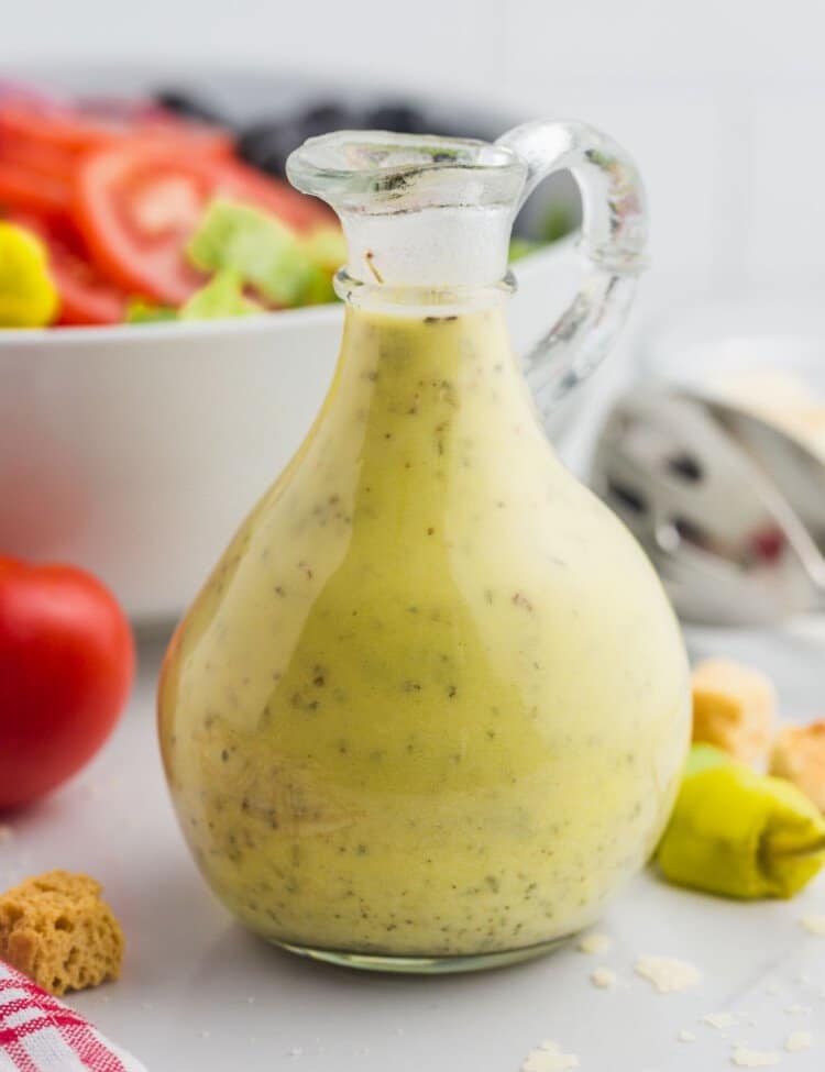 Olive Garden salad dressing in a glass jug, with olive garden salad in a bowl in the background