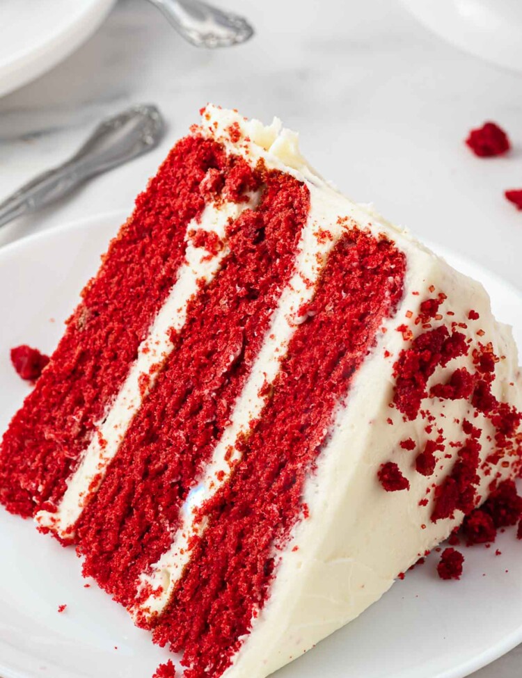 A slice of 3 layer red velvet cake frosted with cream cheese frosting, served on a small white cake plate.