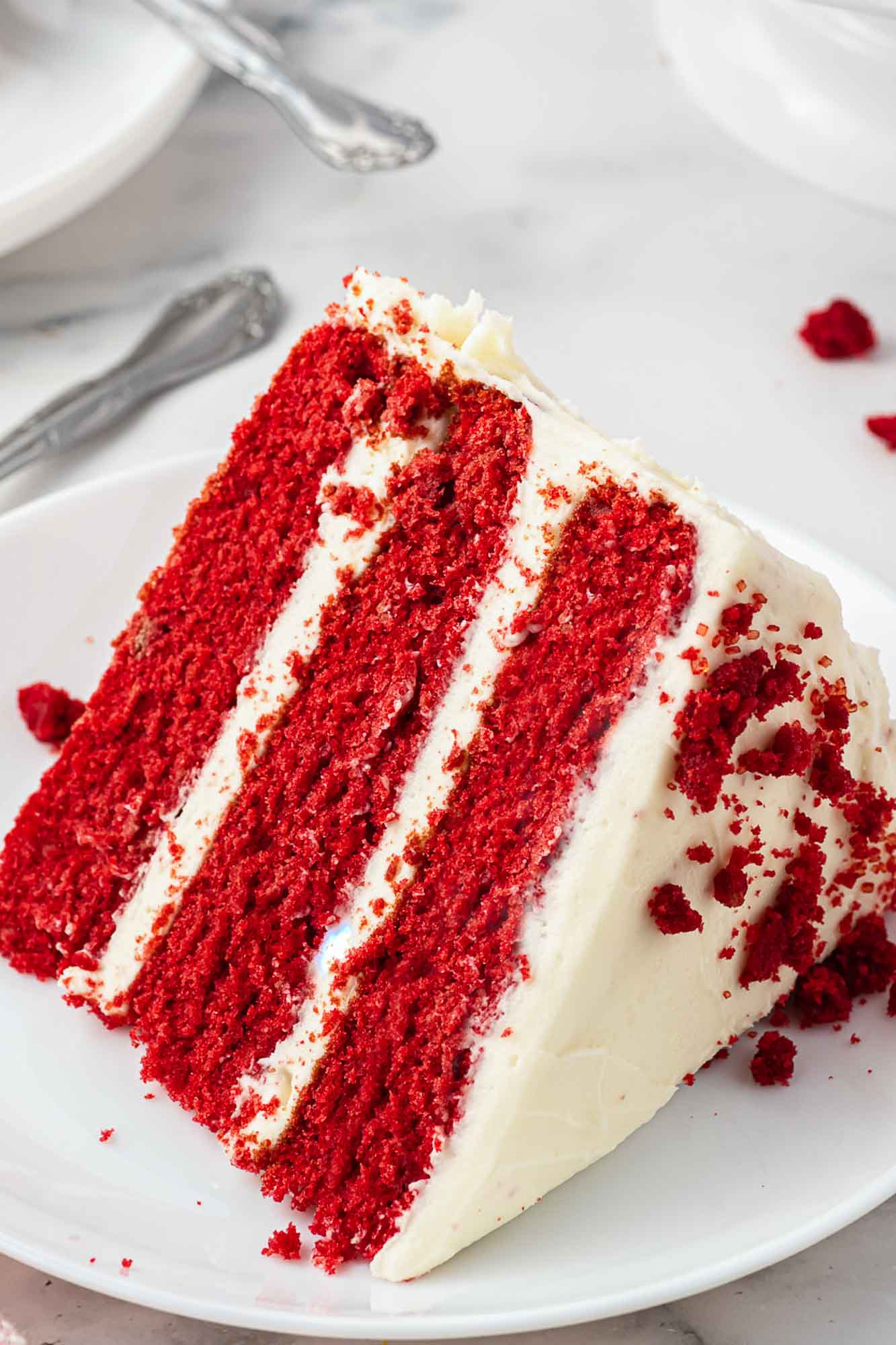 A slice of 3 layer red velvet cake frosted with cream cheese frosting, served on a small white cake plate.
