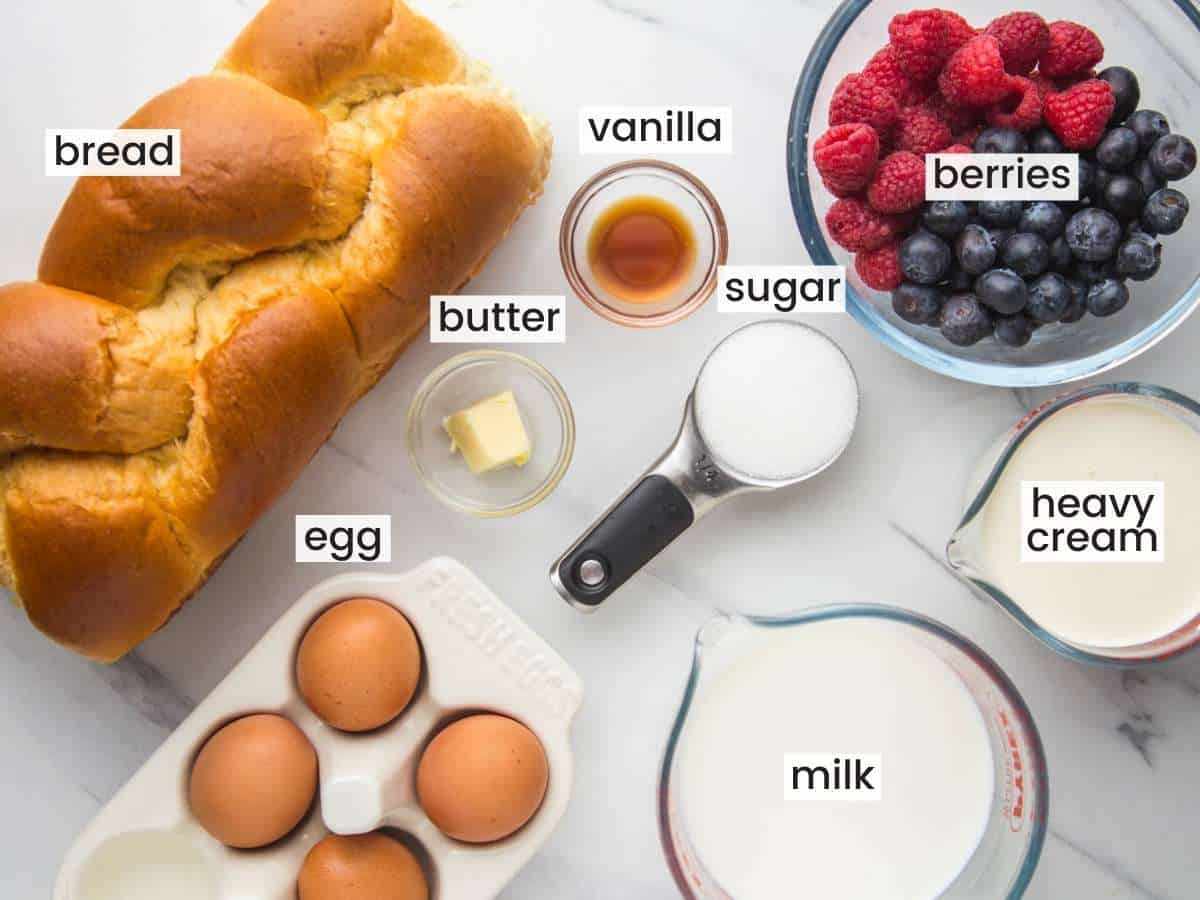 Ingredients needed to make bread pudding including brioche bread, eggs, piece of butter, vanilla, milk, heavy cream, sugar, and fresh berries.