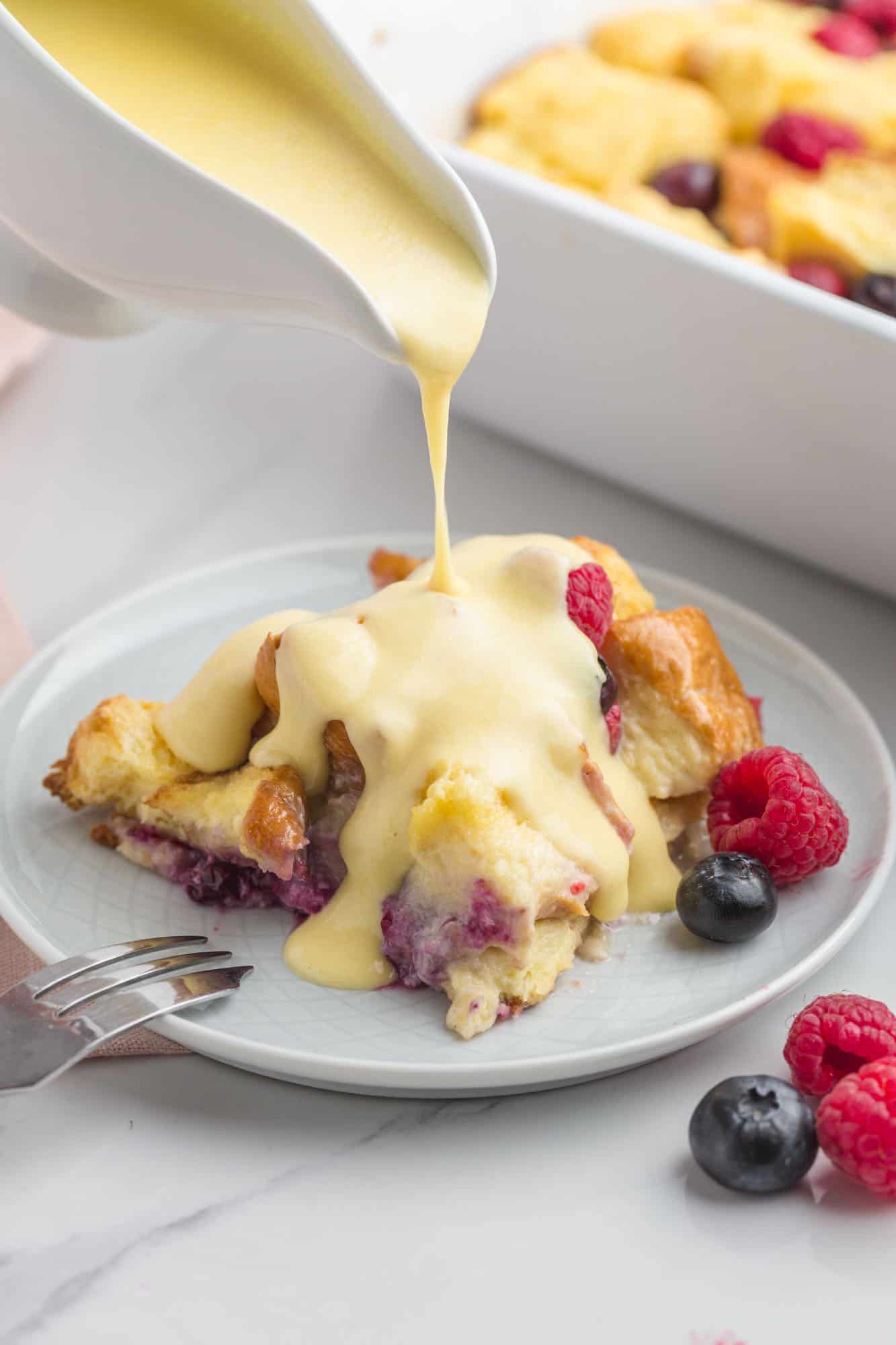 Pouring warm vanilla sauce over a serving of berry bread pudding on a small plate, with fresh berries and a fork on the side.