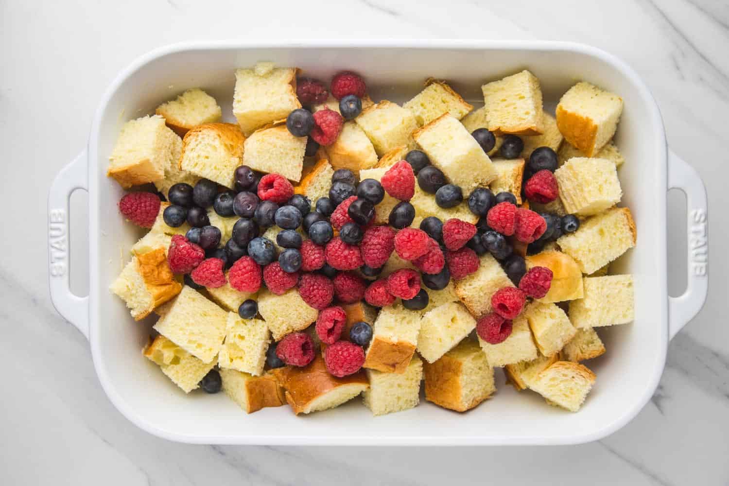 Overhead shot of a white baking dish filled with pieces of bread and fresh raspberries and blueberries