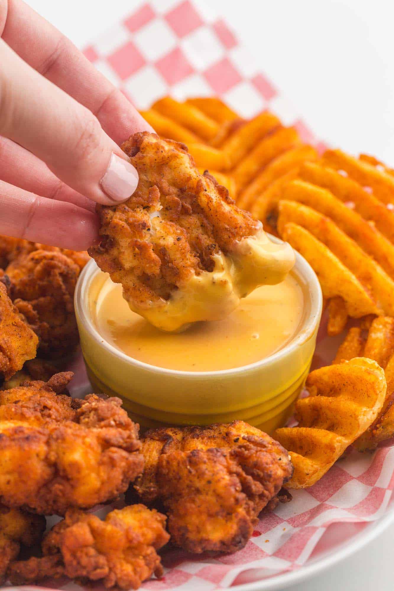 a plate lined with red checkered paper filled with copycat chick fil a nuggets and waffle fries. a hand is dipping a nugget into creamy chick fil a sauce
