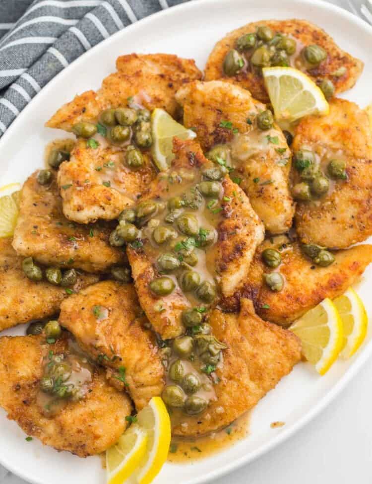 Golden chicken piccata served with lemon caper sauce in a white oval serving dish with fresh lemon slices.