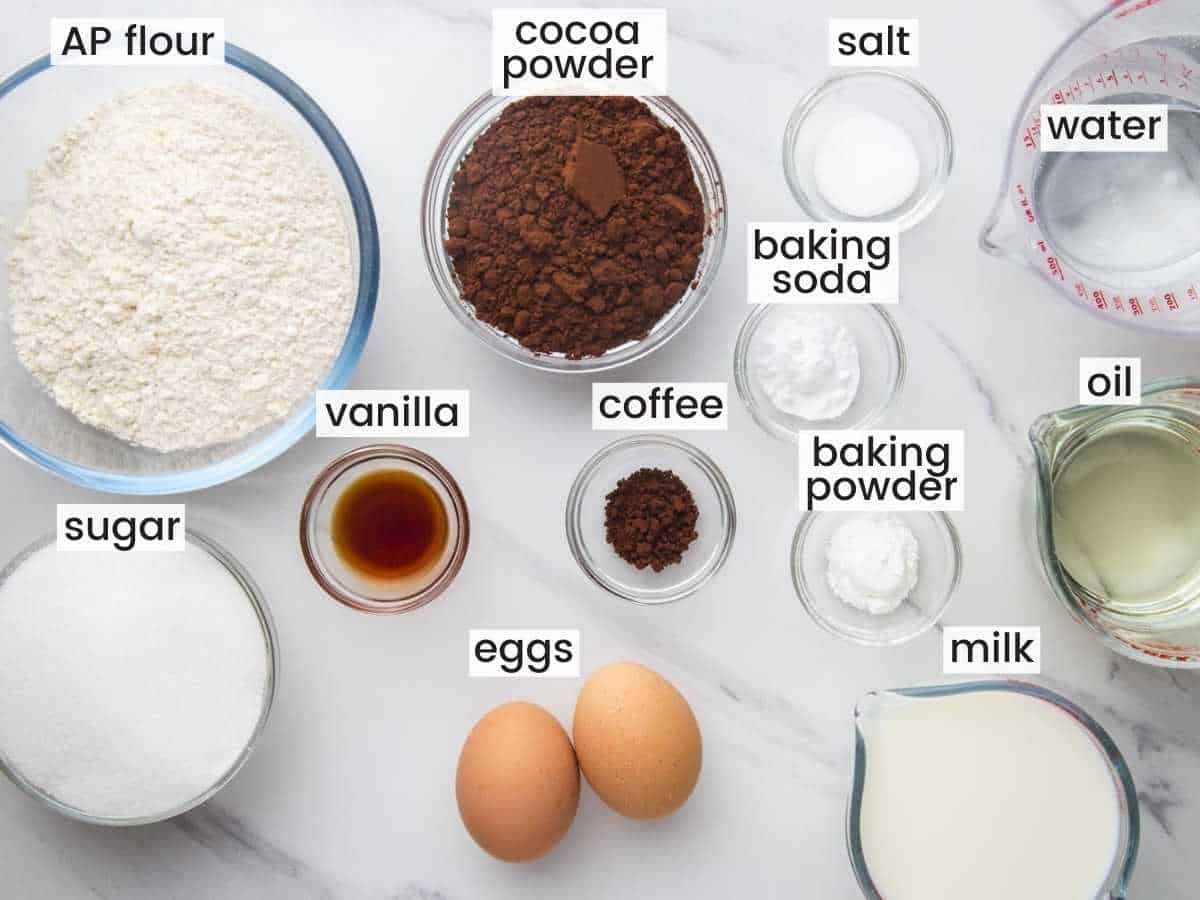 The Ingredients for making a scratch chocolate layer cake, laid out on a marble countertop in separate bowls, viewed from above.