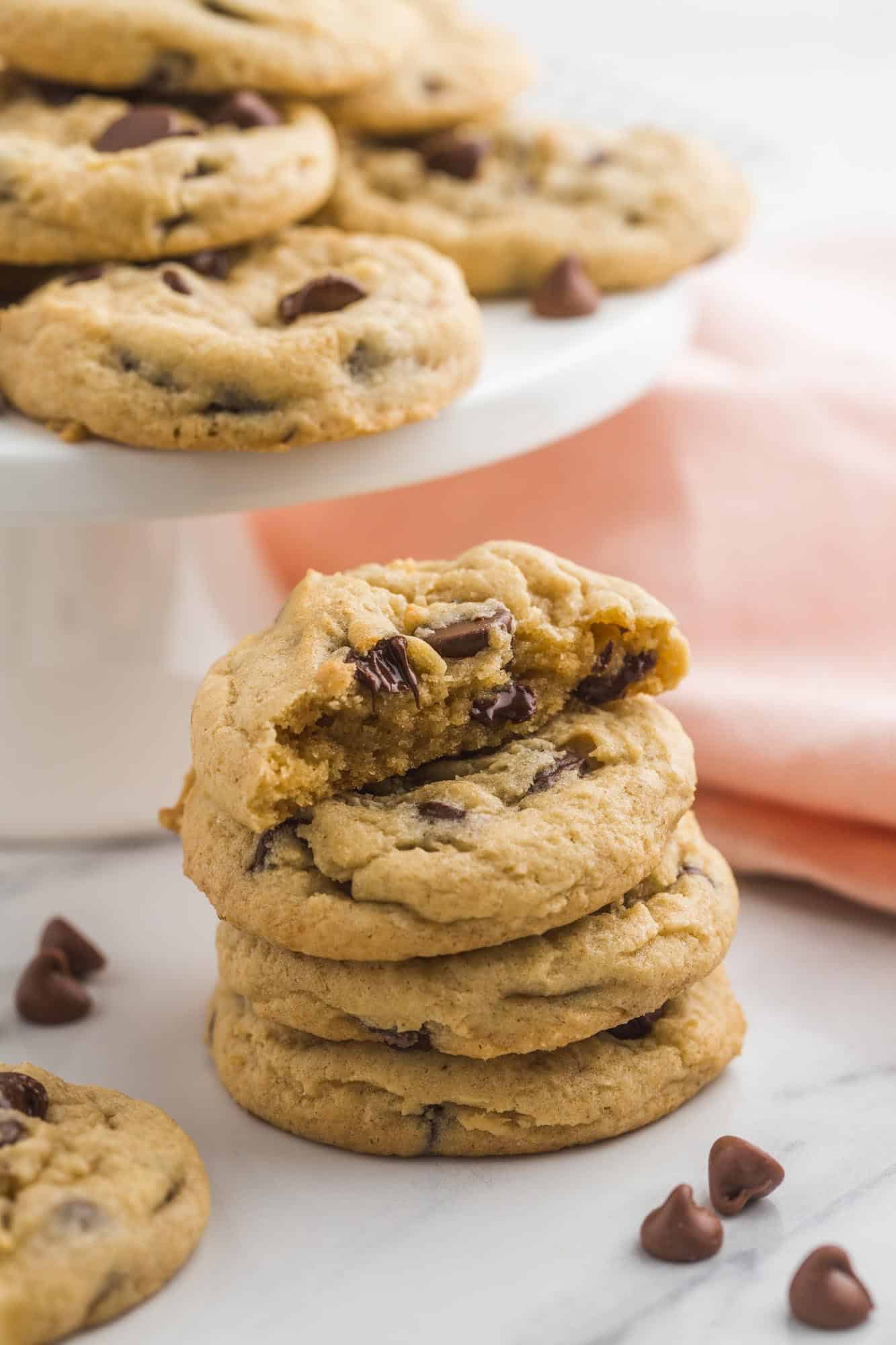 a stack of four chocolate chip pudding cookies on a table in front of a platter of cookies. 