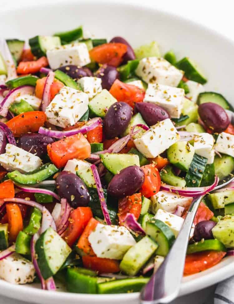 a white bowl filled with chopped greek salad with cucumbers, onions, tomatoes, olives, and feta cheese cubes.