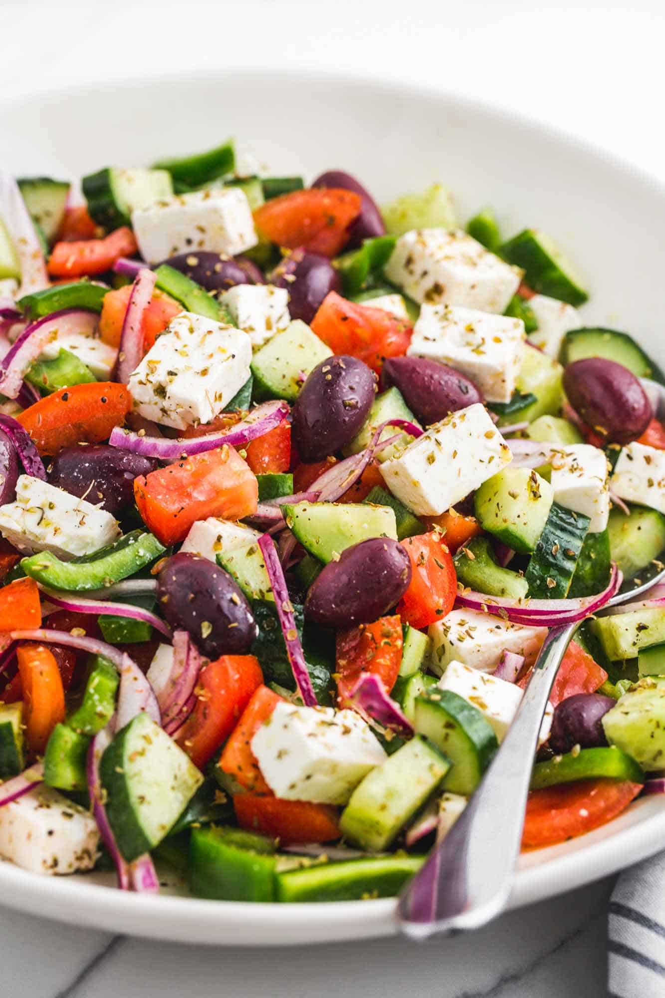 a white bowl filled with chopped greek salad with cucumbers, onions, tomatoes, olives, and feta cheese cubes.