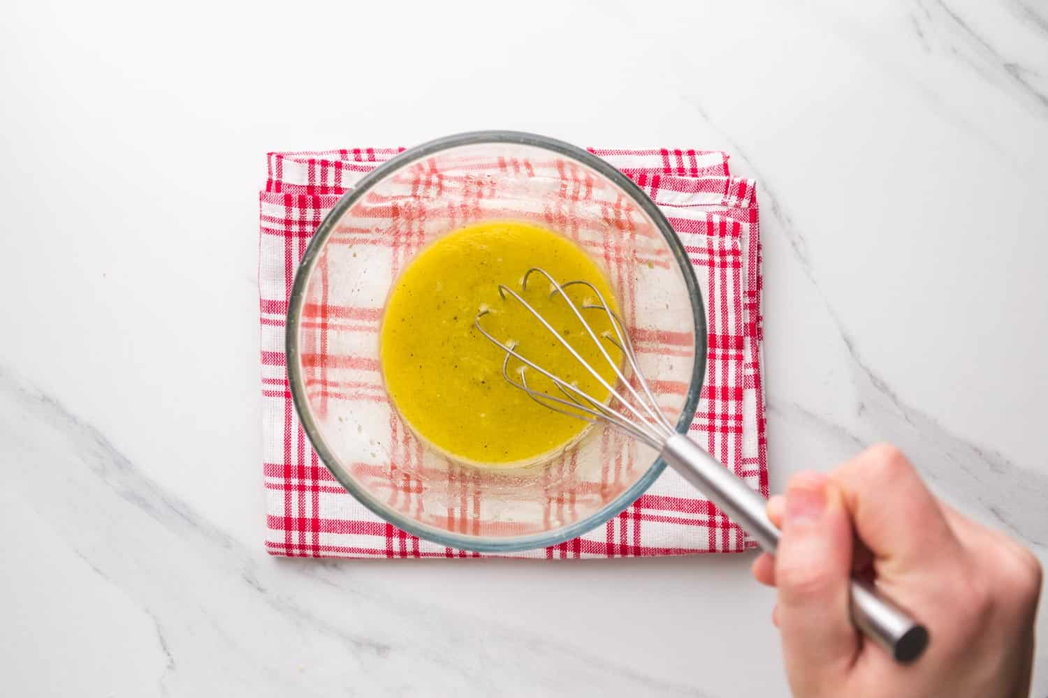 a clear bowl of vinaigrette on a red checked towel, being whisked by hand.