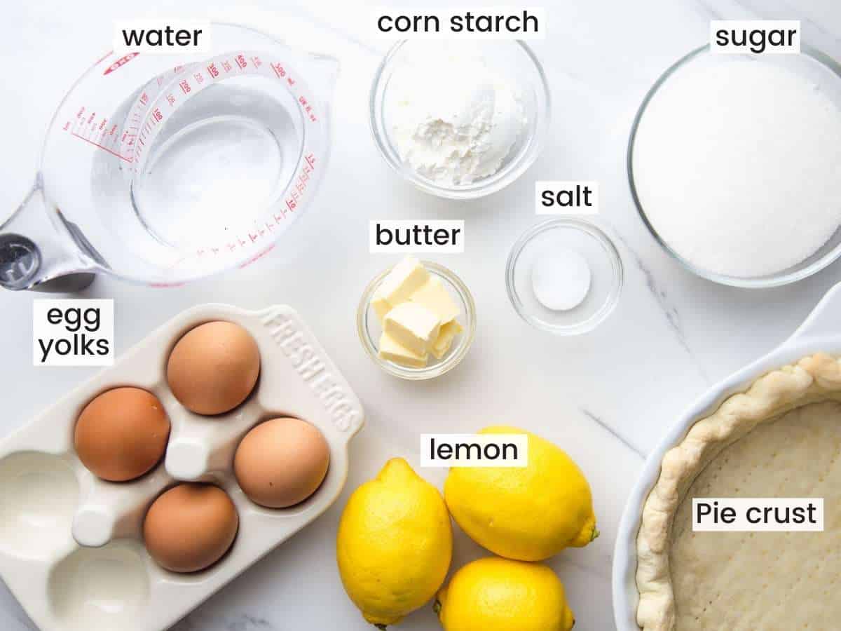 The ingredients needed to make lemon meringue pie on a marble counter top, including a pie crust, fresh lemons, eggs, and sugar.