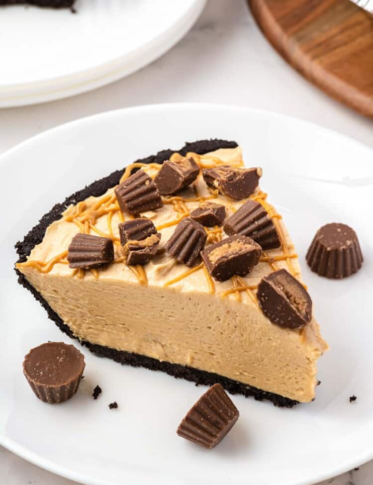 a slice of no bake peanut butter pie with a chocolate crust on a white plate, topped with chopped mini peanut butter cups.