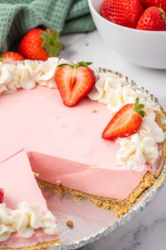 a whole strawberry jello pie in a graham cracker crust. The edge of the pie is decorated with whipped cream and halved strawberries. A slice has been removed.