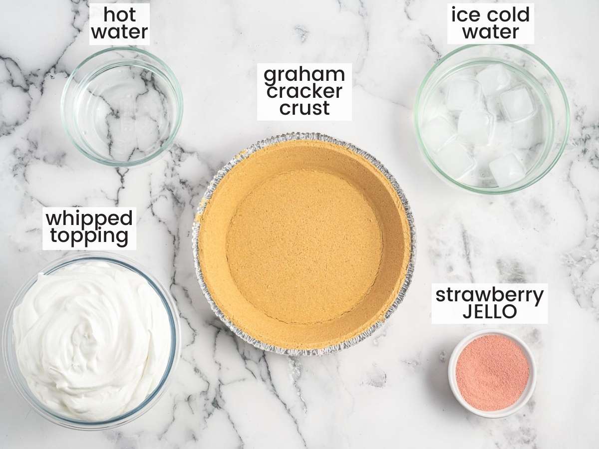 The ingredients for making strawberry no bake pie, measured into bowls on a marbled countertop. A graham cracker crust is in the center, with cool whip, hot water, ice water, and jello powder around it. 