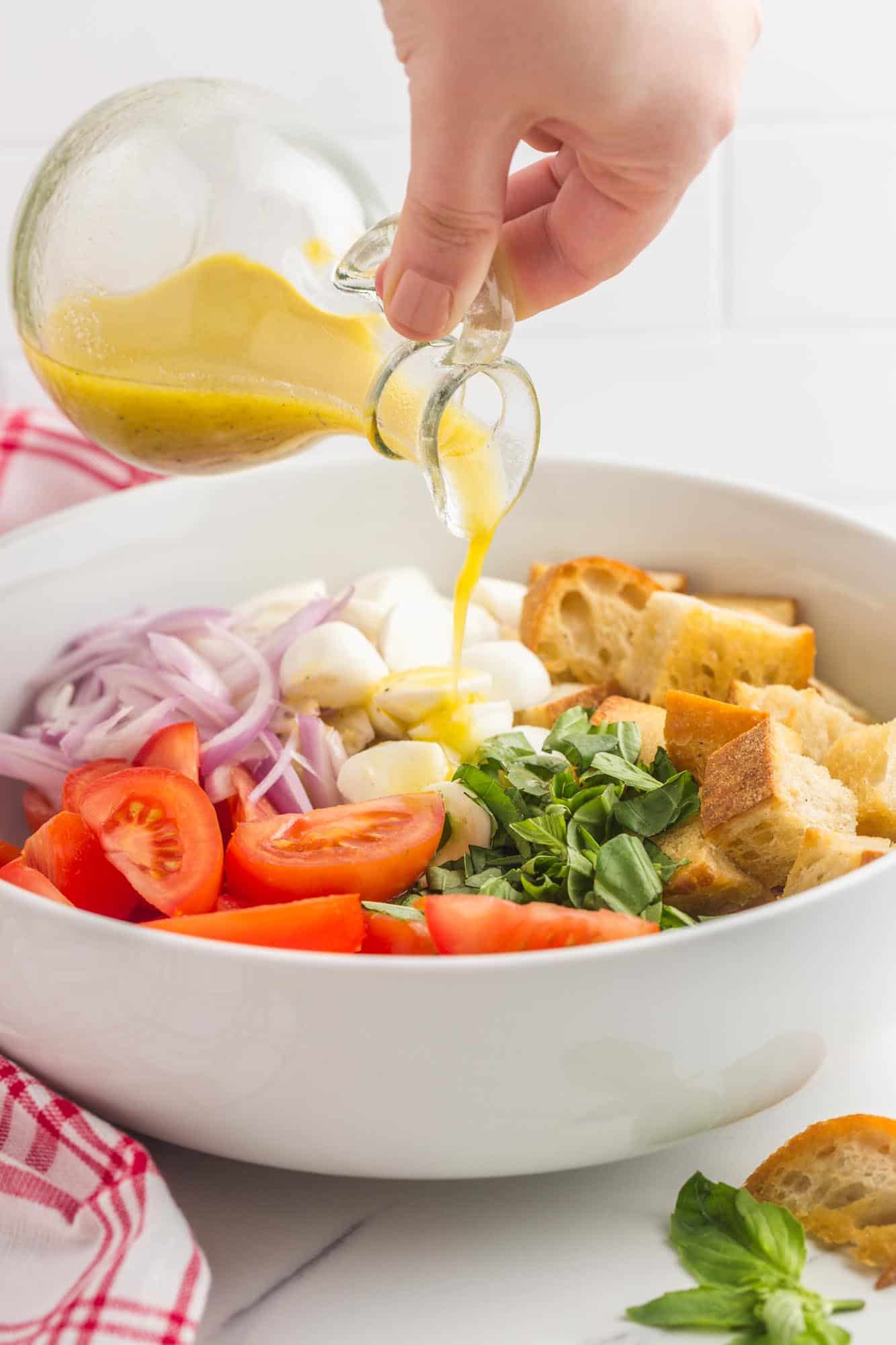 a person's hand pouring dressing onto a panzanella salad in a white bowl.