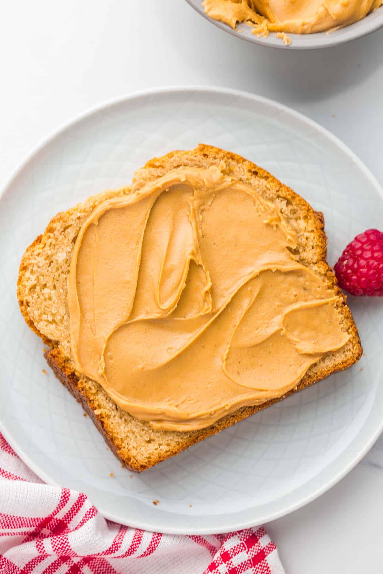Overhead shot of a slice of peanut butter bread with smooth and creamy peanut butter spread on it, and a raspberry on the side.