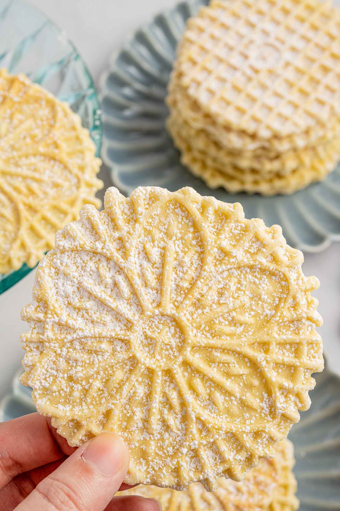 a hand holding a flower pizzelle above blue plates filled with stacks of them