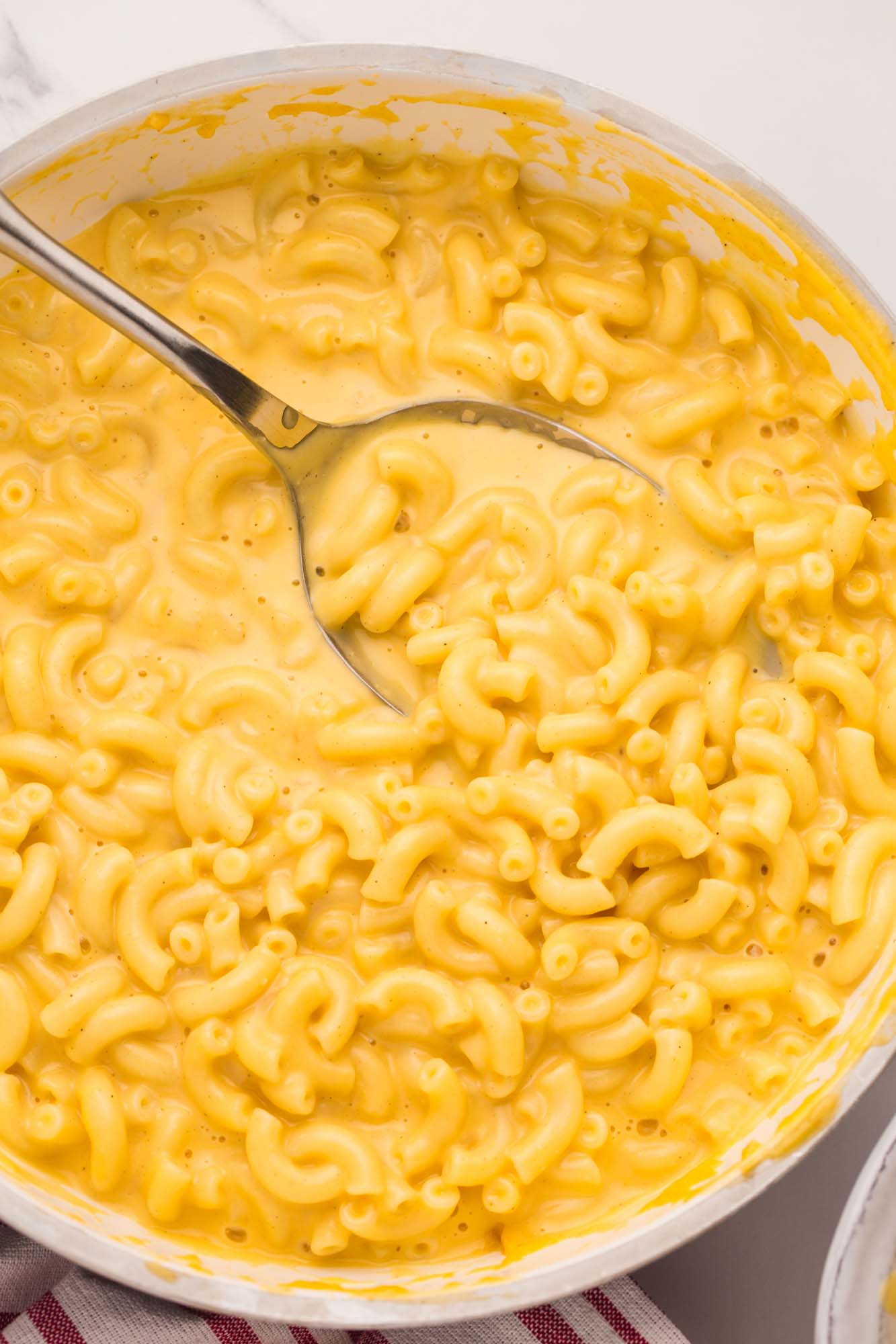 A large white saucepan filled with stovetop mac and cheese made with elbow noodles. a metal serving spoon is stirring it.