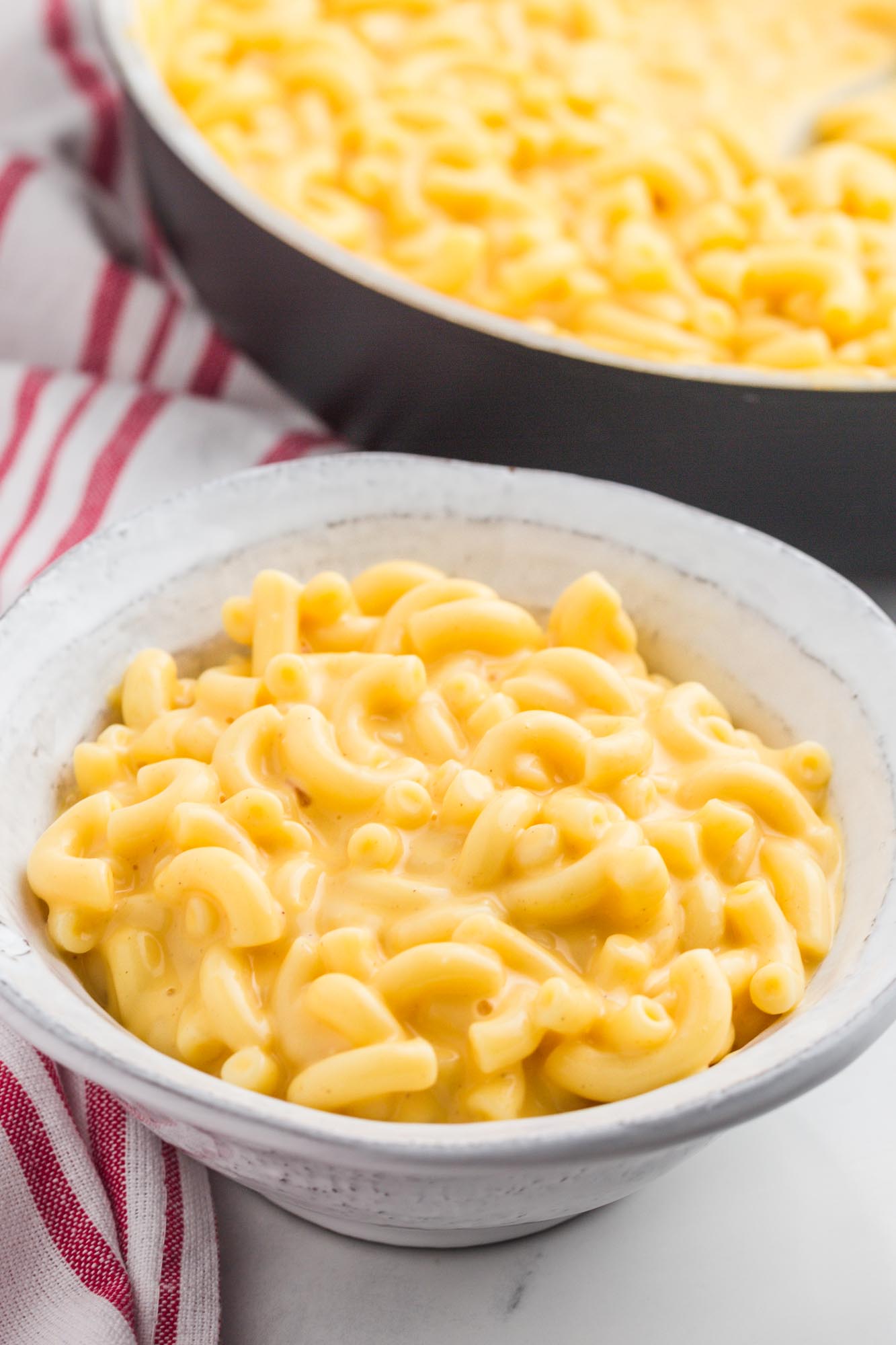 a small white bowl of mac and cheese in front of a skillet of the same. 