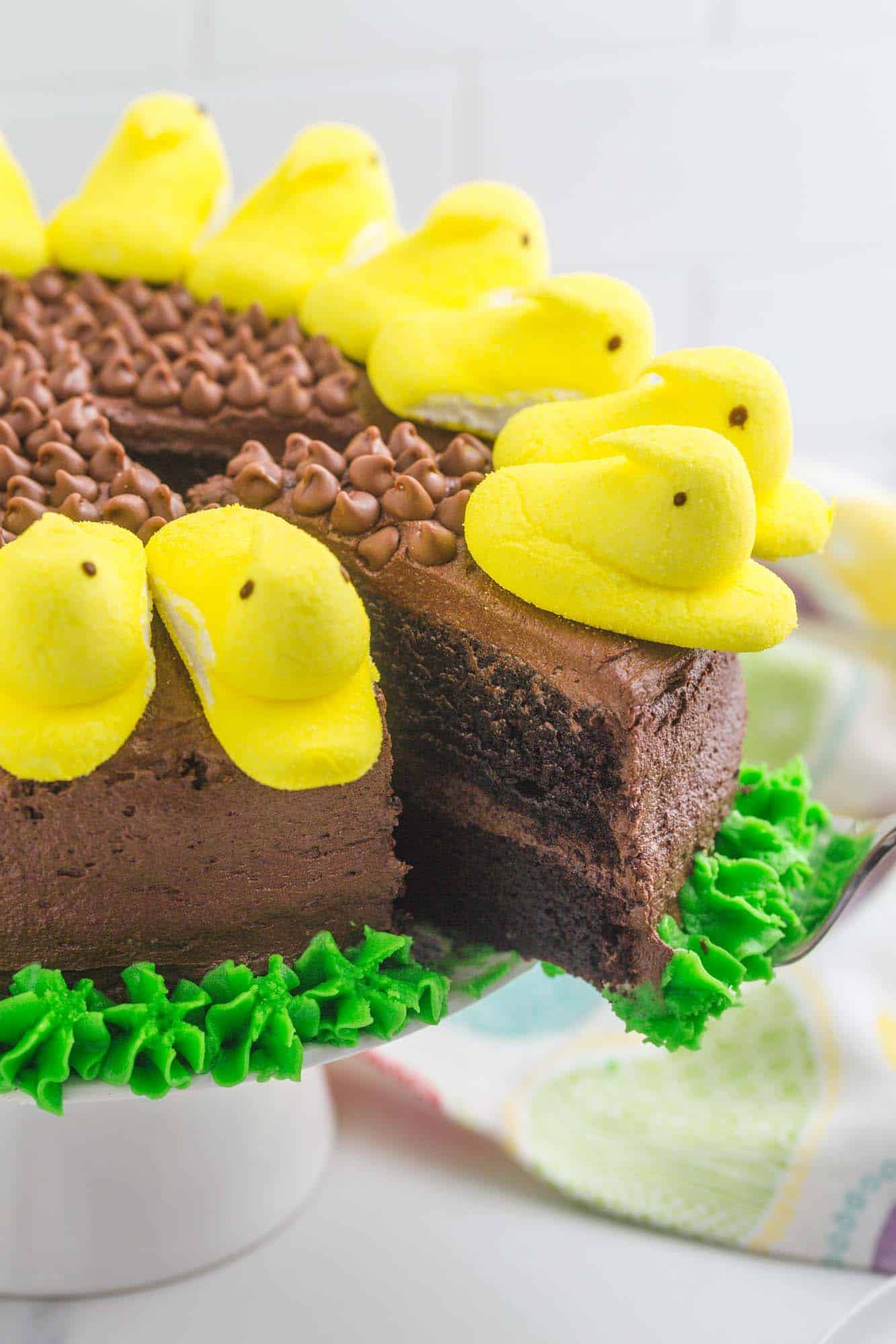 a slice of chocolate cake with yellow peeps on it is being removed from the whole sunflower peeps cake with a cake server.