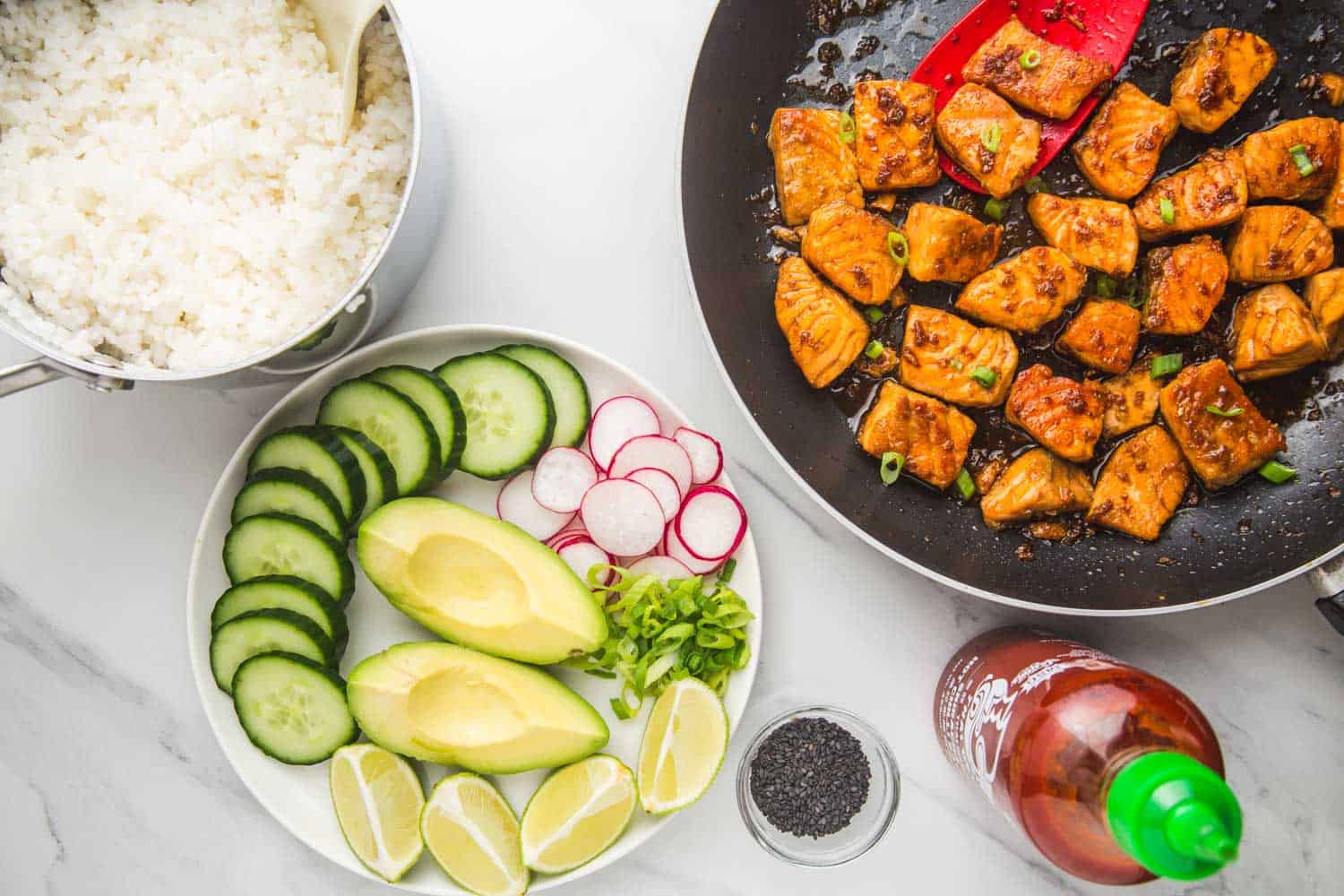 a photo showing how to assemble a salmon rice bowl. The cooked salmon, cooked rice, and sliced vegetables are on a cutting board with sriracha sauce and poppy seeds