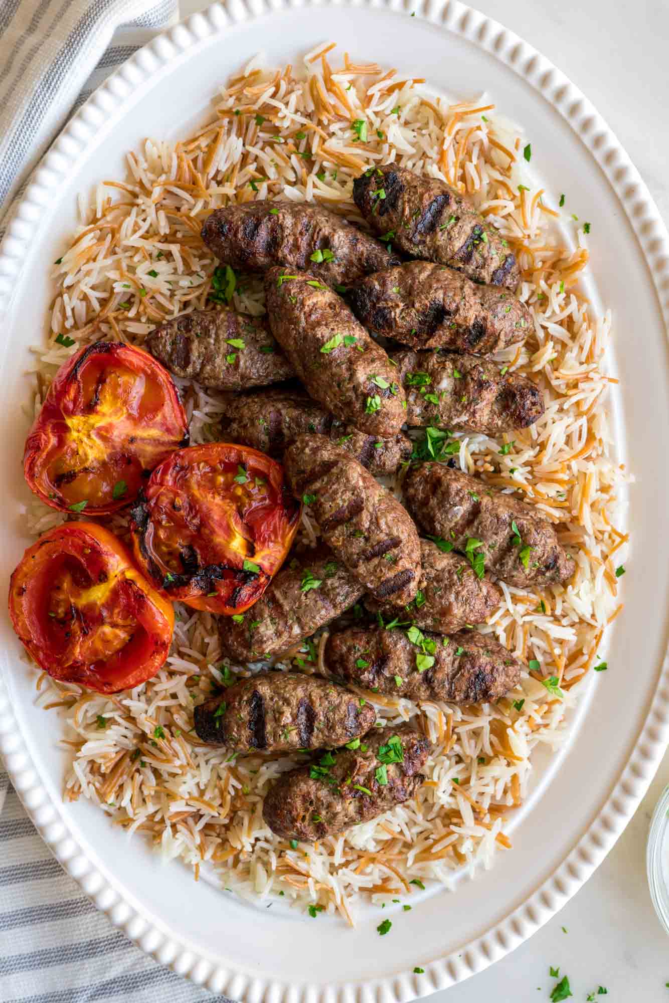 Overhead shot of a large white oval platter with lebanese vermicelli rice, kaftas, and grilled tomatoes.