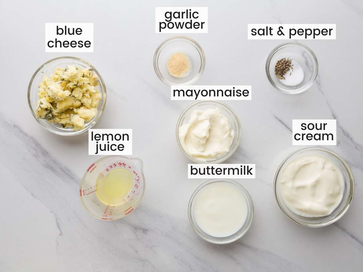 the ingredients for making a homemade blue cheese dressing in small bowls on a marble countertop