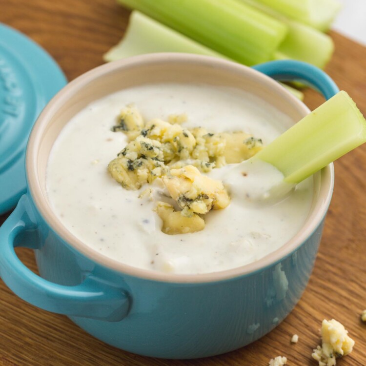 a blue crock filled with blue cheese dressing. a stack of celery sticks is in the background, and one celery stick is in the blue cheese.